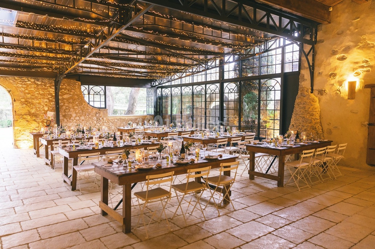 Salle de réception lumineuse avec tables en bois, chaises blanches et guirlandes suspendues sous un toit en verrière.