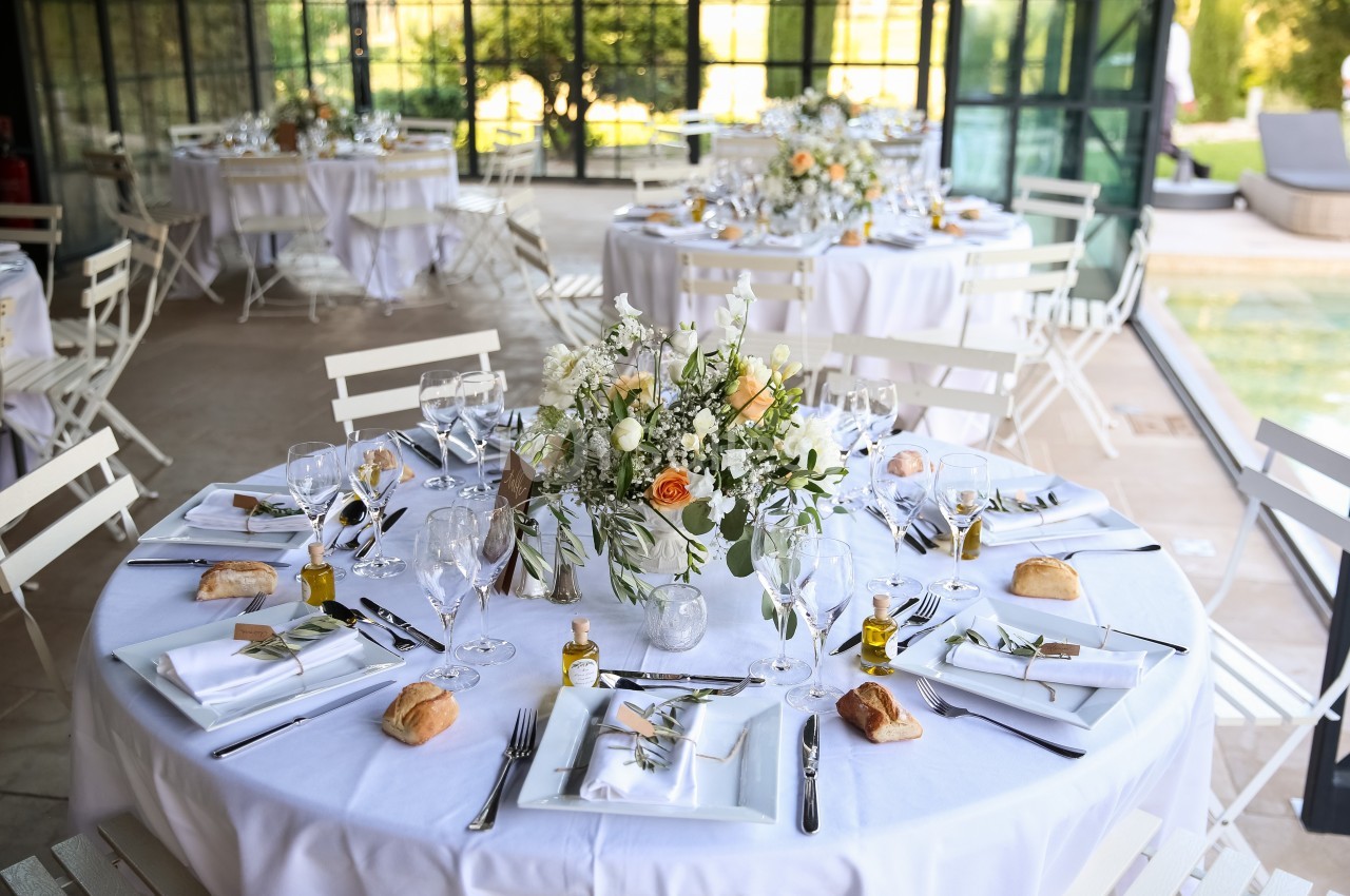Table ronde élégamment dressée avec nappes blanches, vaisselle raffinée, bouquets floraux et petits pains individuels.