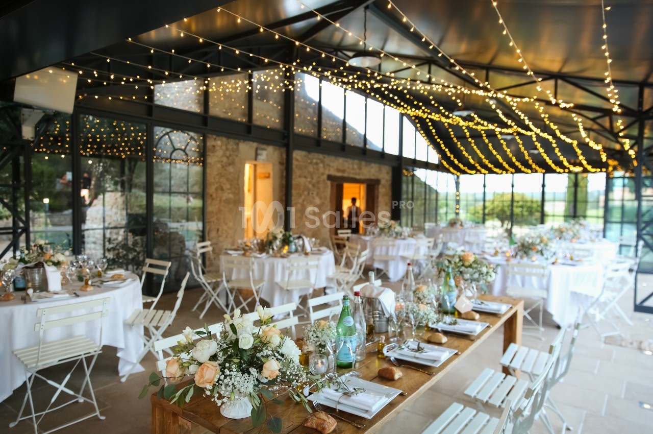 Salle de réception lumineuse décorée pour un mariage, avec tables dressées et guirlandes lumineuses suspendues.