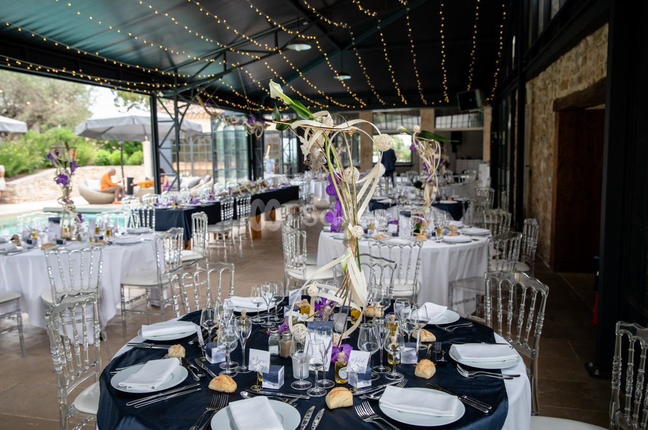 Salle de réception décorée pour un événement, avec tables rondes dressées, nappes sombres et centres de table fleuris.