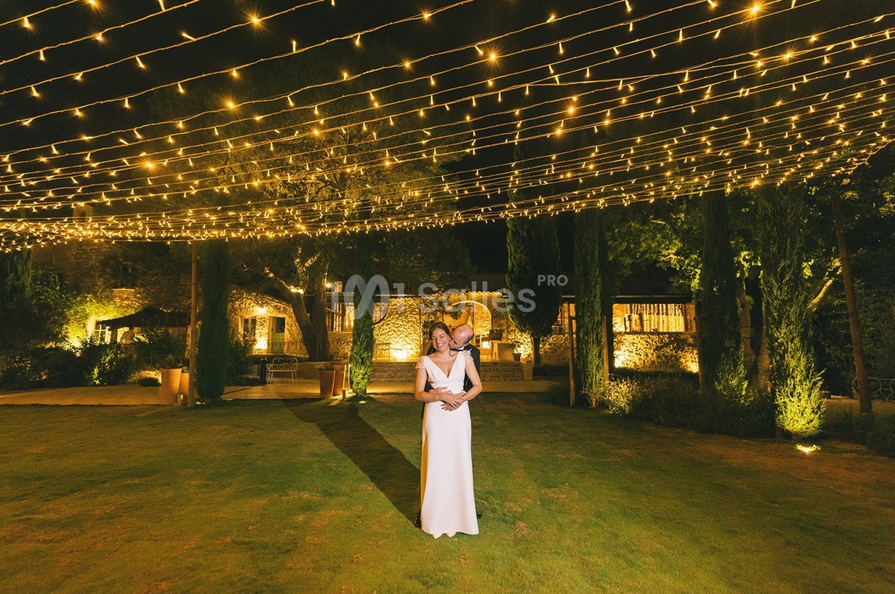 Un couple pose sous des guirlandes lumineuses dans un jardin éclairé, entouré de végétation et de bâtiments en pierre.