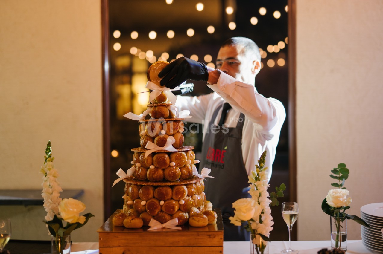 Un serveur assemble une pièce montée de choux décorée de rubans blancs, entourée de fleurs et de lumières.