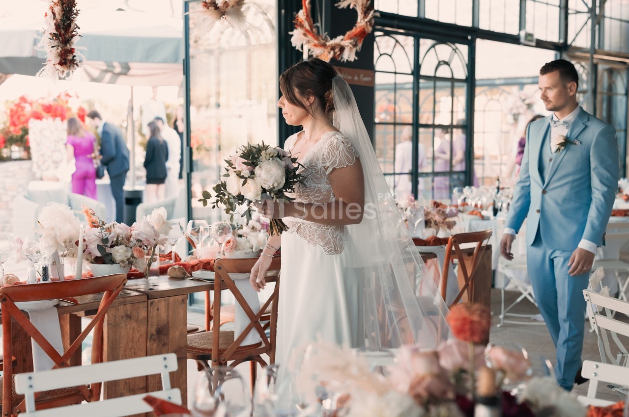 Une mariée en robe blanche tient un bouquet dans une salle décorée pour un mariage, avec un homme en costume bleu derrière…