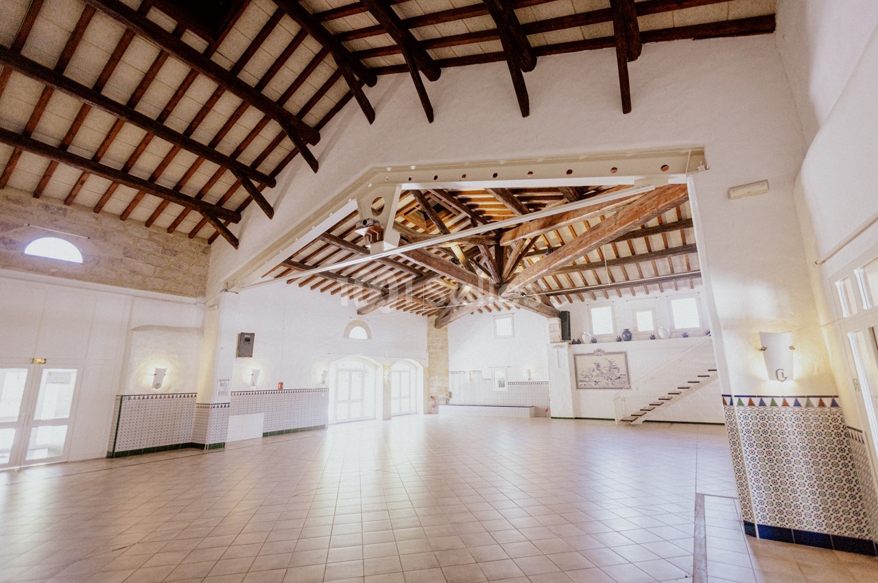 Grande salle lumineuse avec plafond en poutres apparentes, murs blancs, carrelage au sol et escaliers sur le côté.