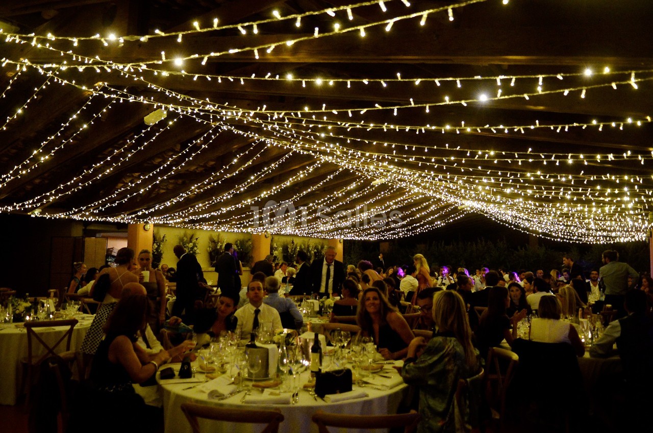 Salle de réception décorée de guirlandes lumineuses, avec des invités assis à des tables rondes.