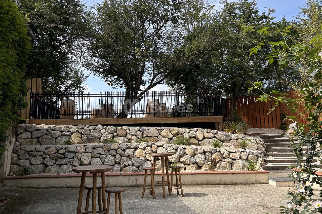 Terrasse surélevée entourée d'arbres, avec des tables en bois sur un sol pavé et un muret en pierres en premier plan.