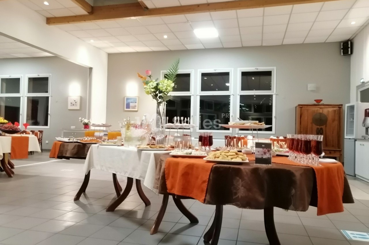 Buffet dressé dans une salle lumineuse avec nappes blanches et orange, proposant boissons et plats variés.