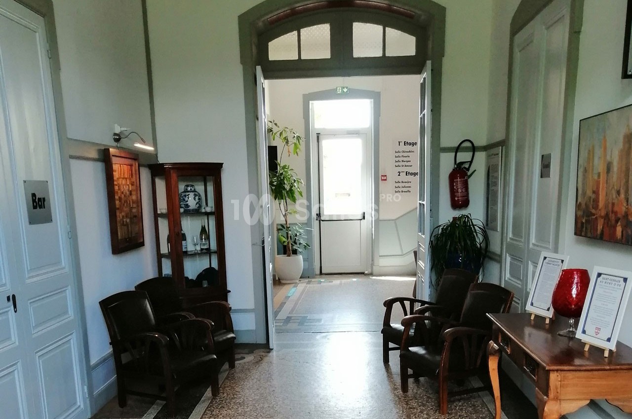 Un couloir lumineux avec des fauteuils, une armoire vitrée, une table et une porte ouverte sur l'extérieur.