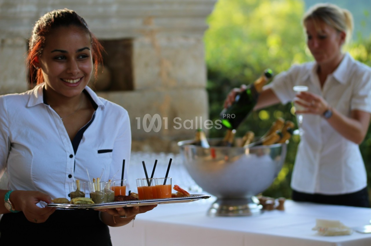 Une serveuse souriante porte un plateau de boissons, tandis qu'une autre personne sert du champagne en arrière-plan.