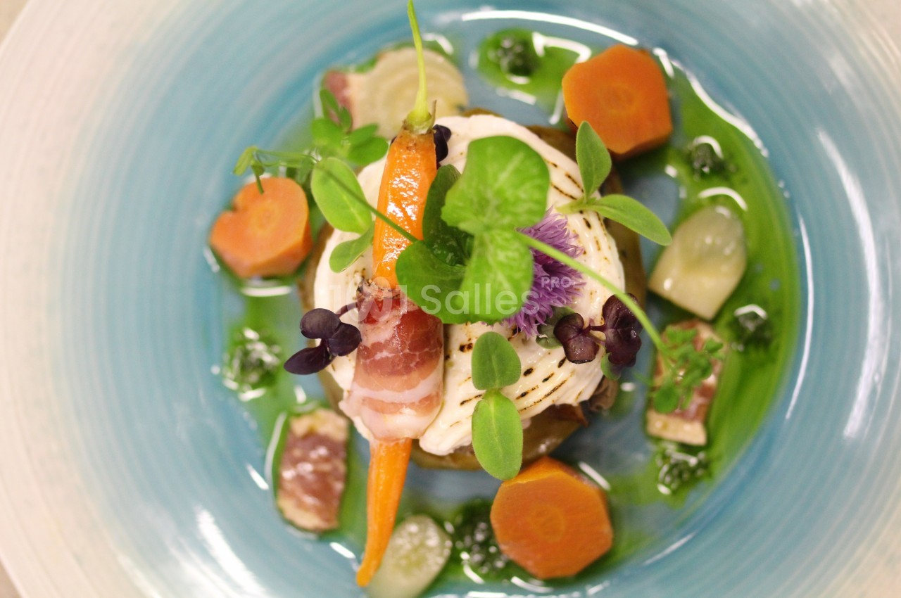 Assiette gastronomique avec légumes colorés, viande, herbes fraîches et sauce verte, présentée sur une assiette bleue.