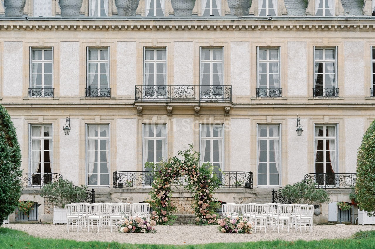 Façade d'un bâtiment élégant avec des chaises blanches disposées devant une arche florale pour une cérémonie en extérieur.