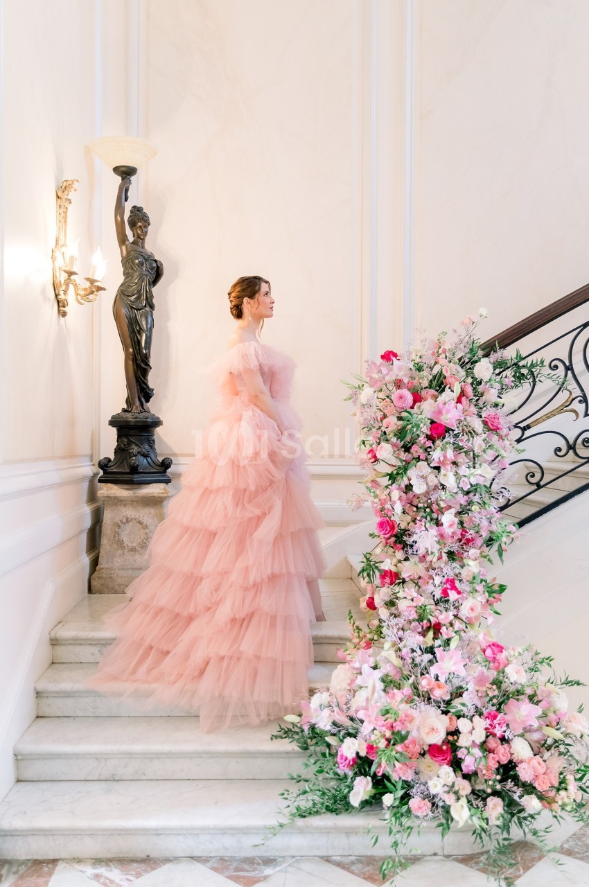 Femme en robe rose volumineuse montant un escalier décoré de fleurs colorées, dans un intérieur élégant.