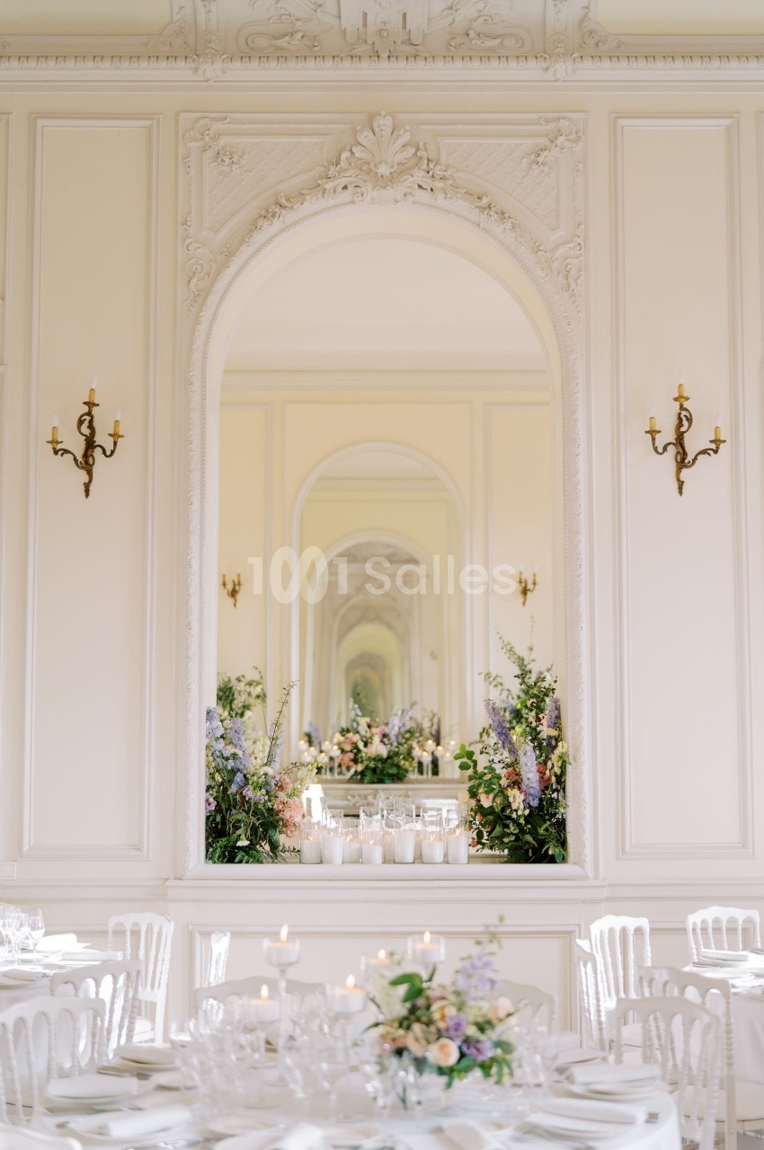 Salle élégante avec moulures, miroir central reflétant des compositions florales, tables dressées et chaises blanches.