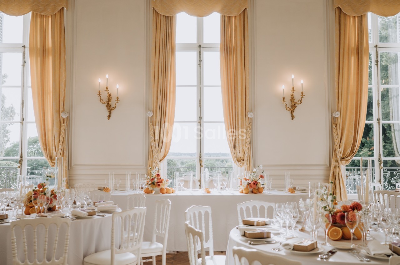 Salle de réception élégante avec tables dressées, nappes blanches, décorations florales et grandes fenêtres encadrées de…