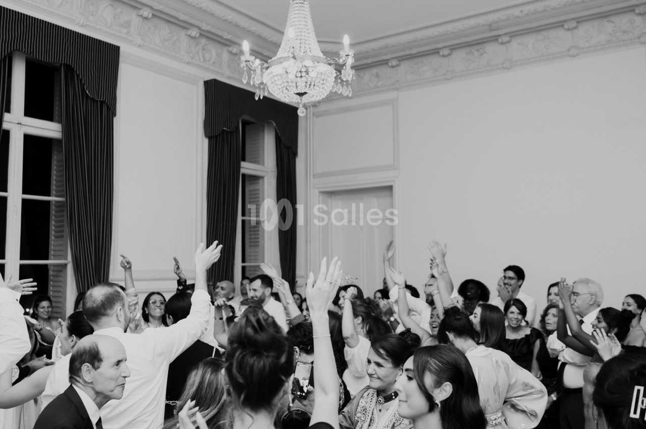 Des personnes dansent et lèvent les bras lors d'une fête dans une salle élégante avec un lustre au plafond.