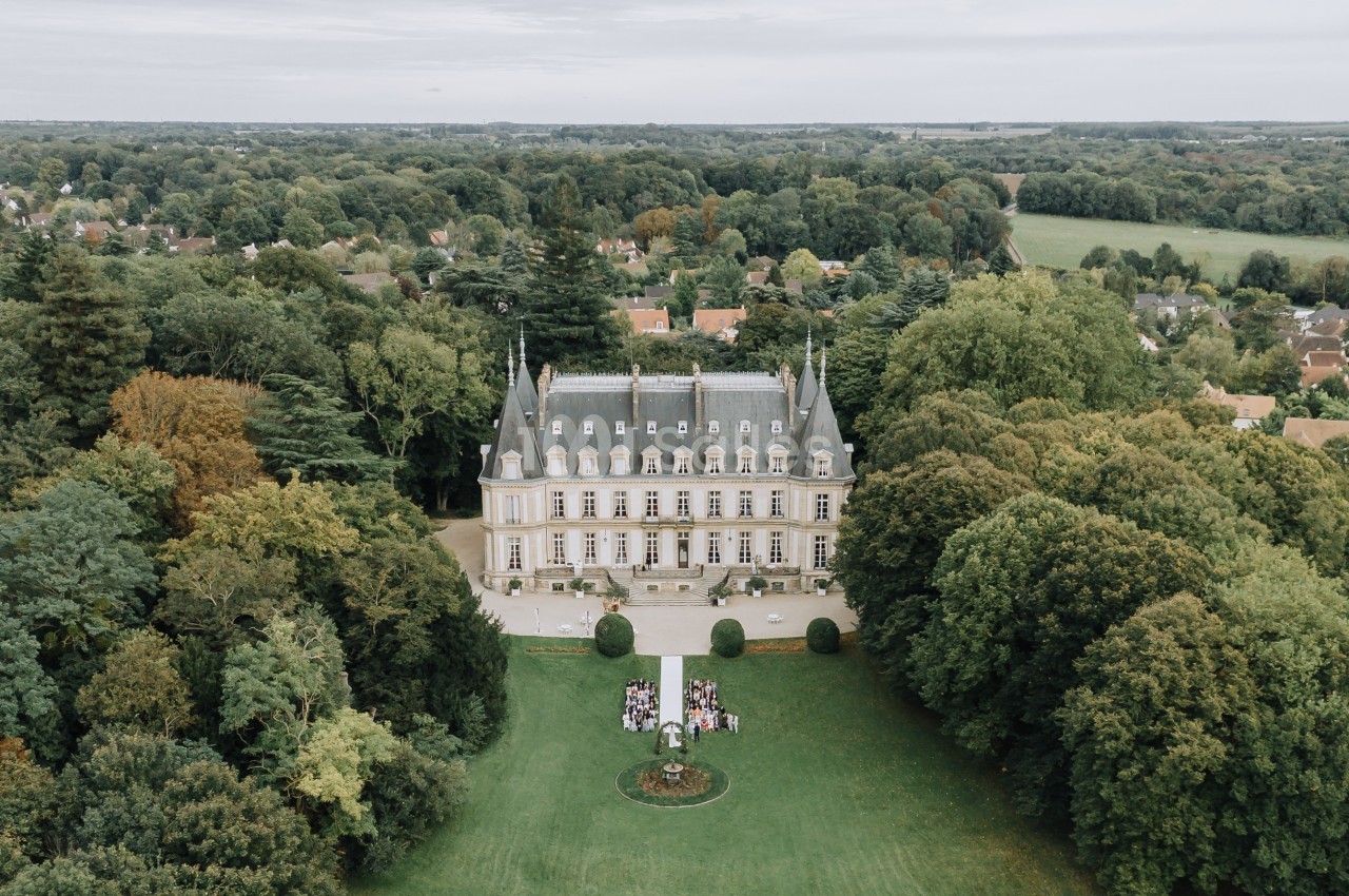 Vue aérienne d'un château entouré de jardins et d'arbres, avec une cérémonie en plein air sur la pelouse.