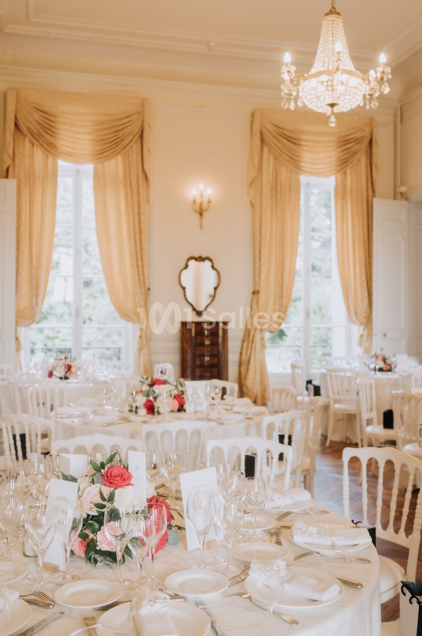Salle de réception élégante avec tables dressées, nappes blanches, décorations florales et lustre suspendu.