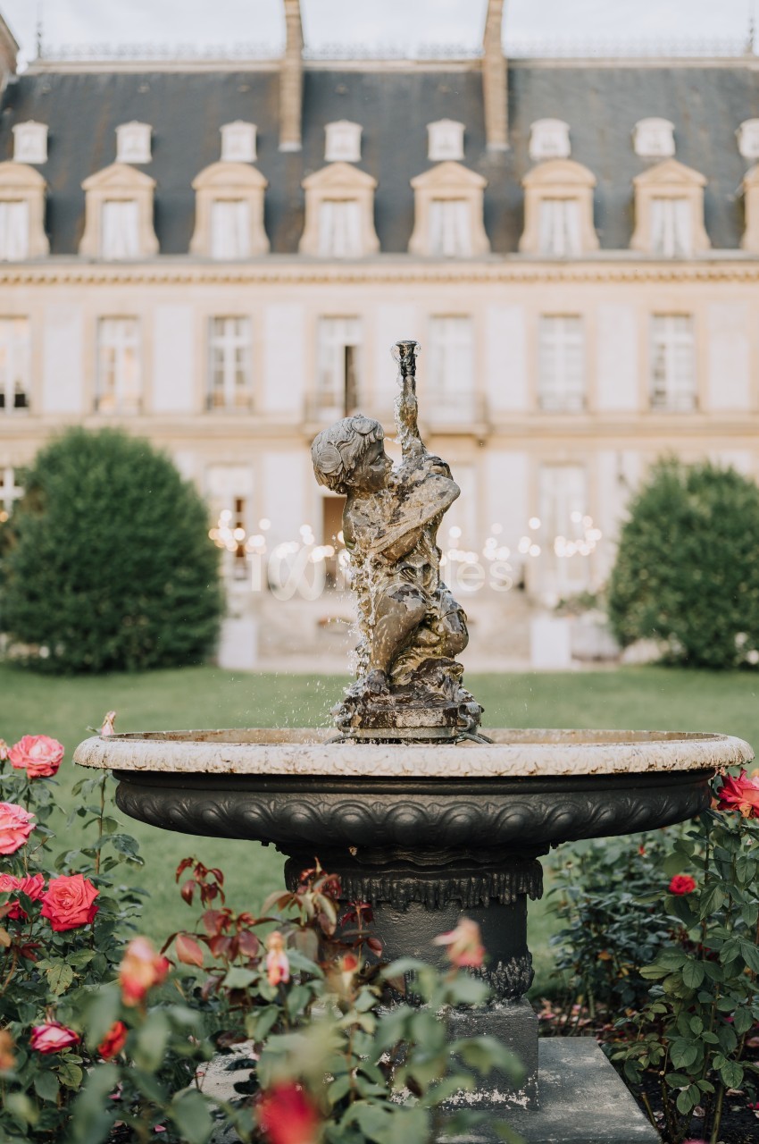 Fontaine ornée d'une sculpture au centre d'un jardin fleuri, avec un bâtiment historique en arrière-plan.