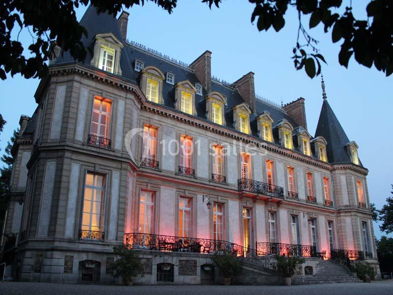 Façade d'un château éclairé par des lumières colorées au crépuscule, entouré de végétation.