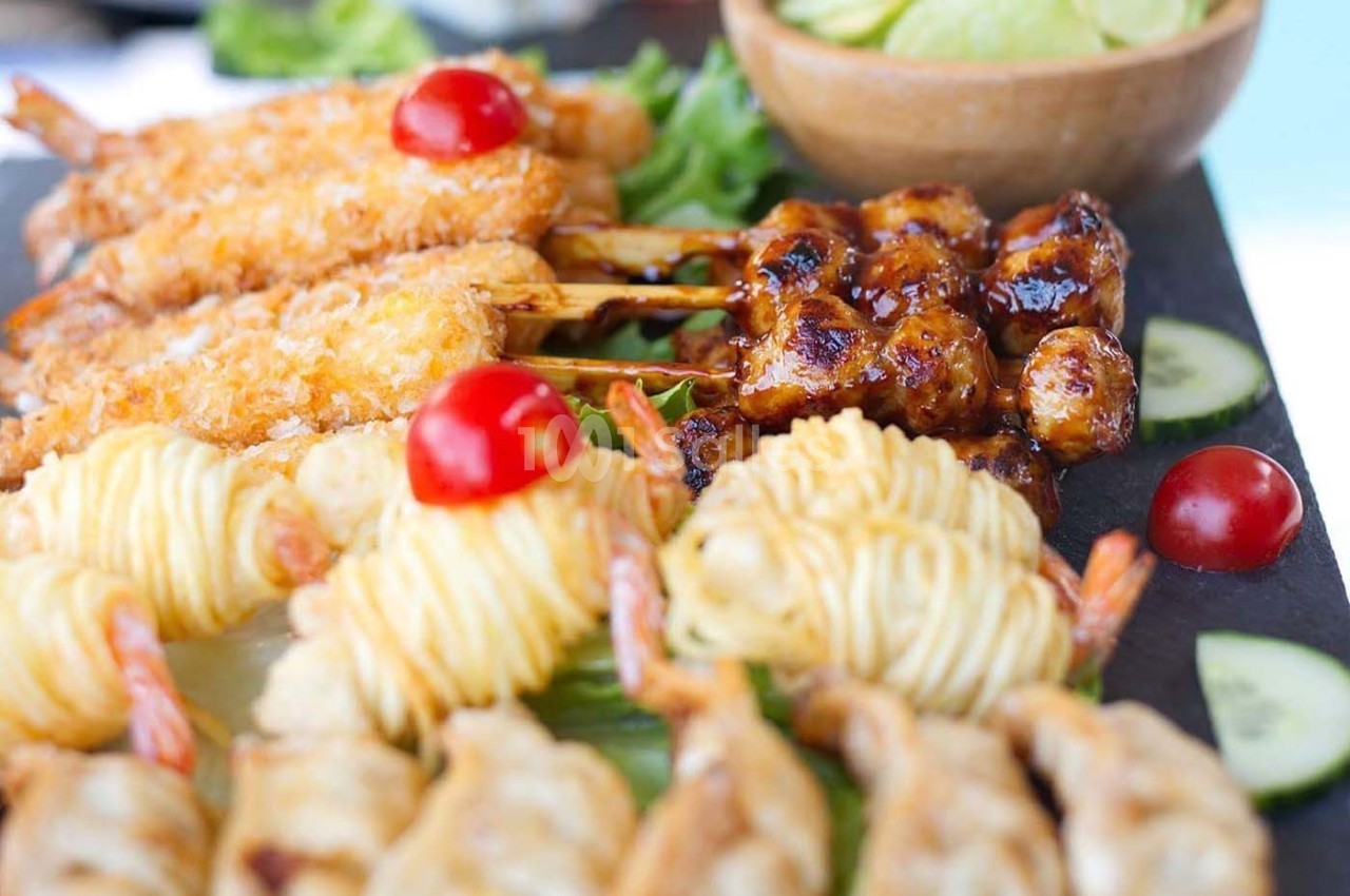 Assortiment de brochettes et bouchées apéritives avec crevettes panées, nouilles et légumes en arrière-plan.