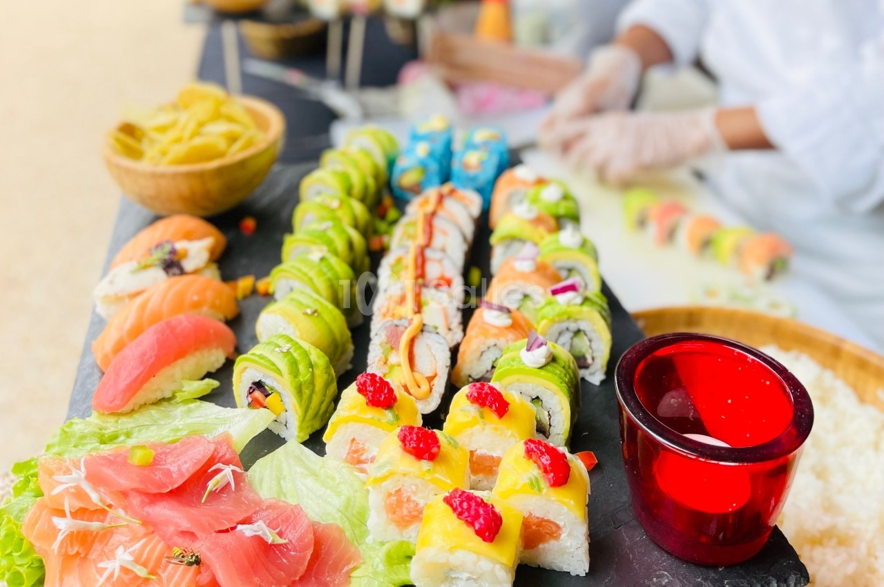 Plateau de sushis variés colorés avec un chef en arrière-plan préparant des rouleaux sur une table.