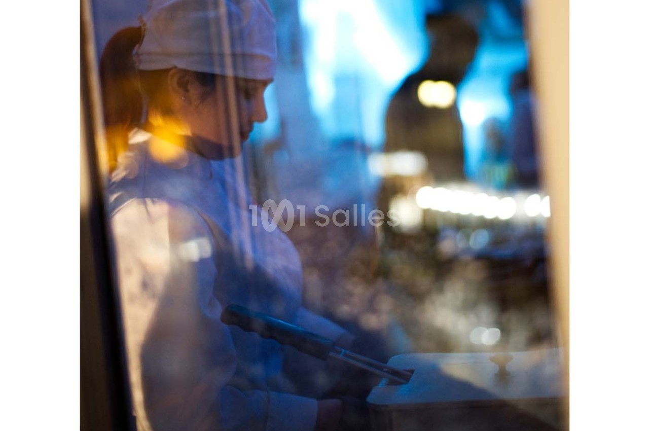 Une cuisinière en uniforme blanc, vue à travers une vitre, prépare un plat dans un environnement éclairé.