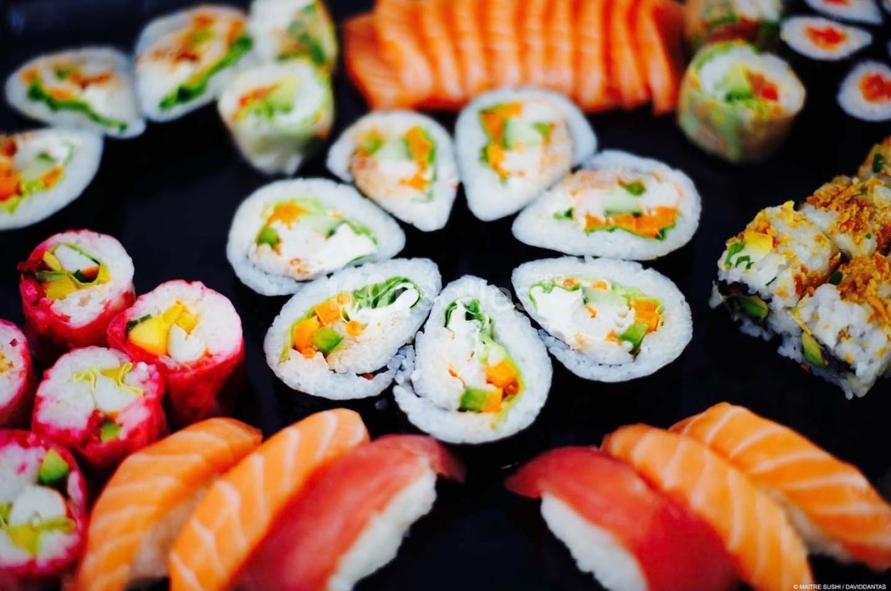 Assortiment de sushis variés disposés sur une surface noire, incluant makis, nigiris et rouleaux colorés.