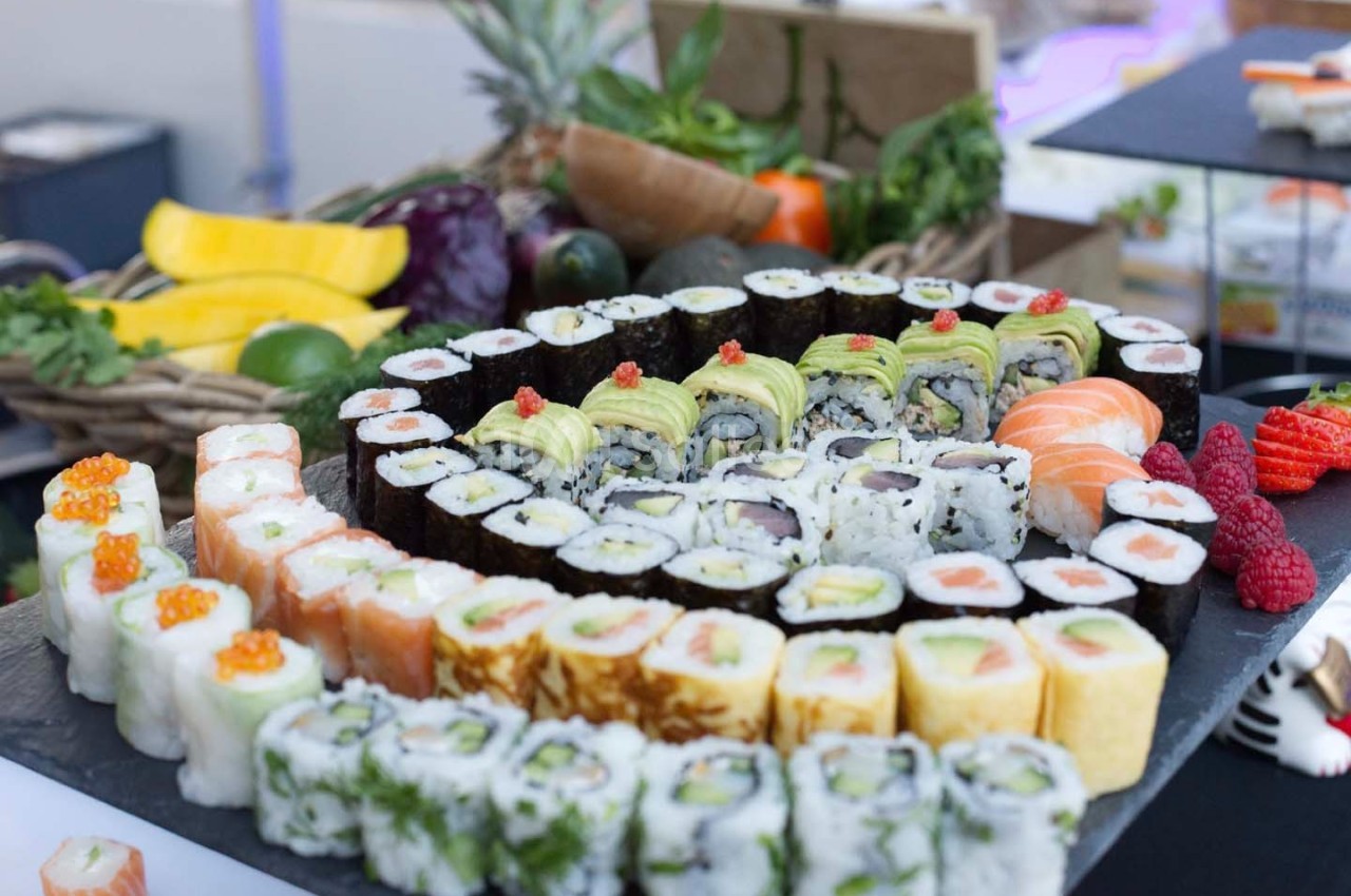 Plateau de sushis variés disposés en cercles sur une ardoise, entouré de fruits et légumes en arrière-plan.