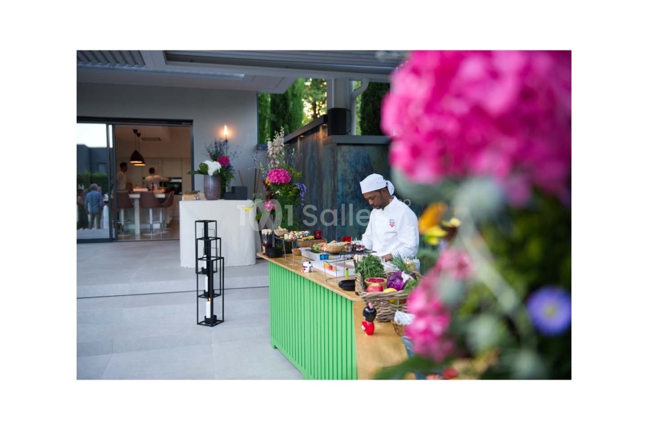 Un chef prépare des plats sur un comptoir extérieur coloré, entouré de fleurs et de verdure.
