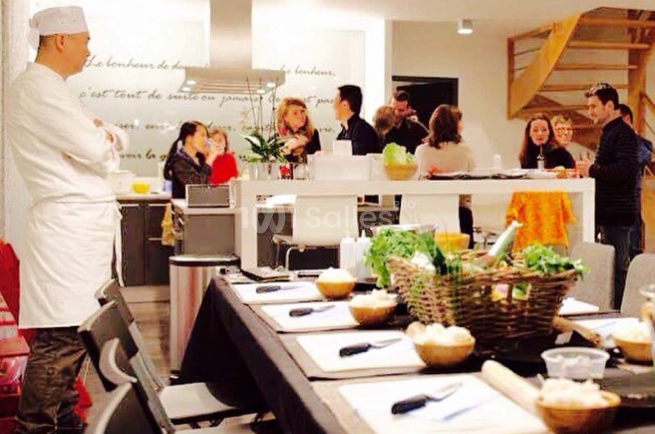 Un chef en tenue blanche observe un groupe de personnes discutant autour d'une table dans une cuisine conviviale.
