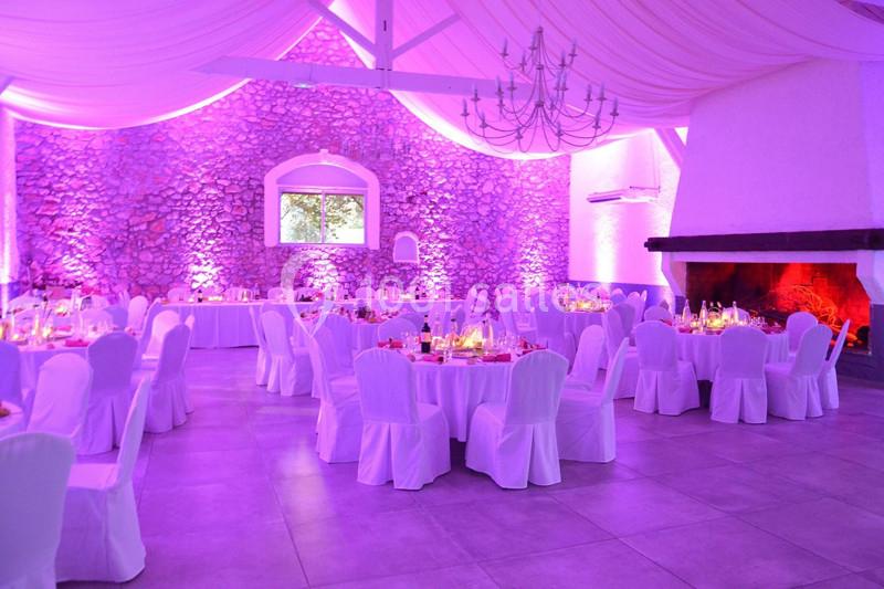 Salle de réception avec murs en pierre, tables dressées, chaises blanches et éclairage rose tamisé.