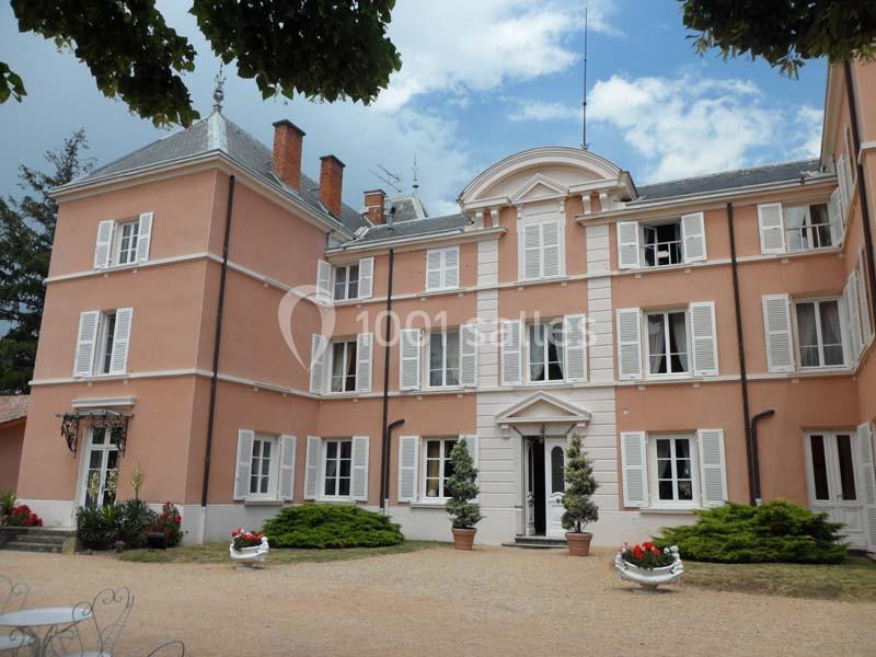 Façade d'un grand bâtiment ancien en crépi rose avec volets blancs, entouré de plantes et d'une cour gravillonnée.