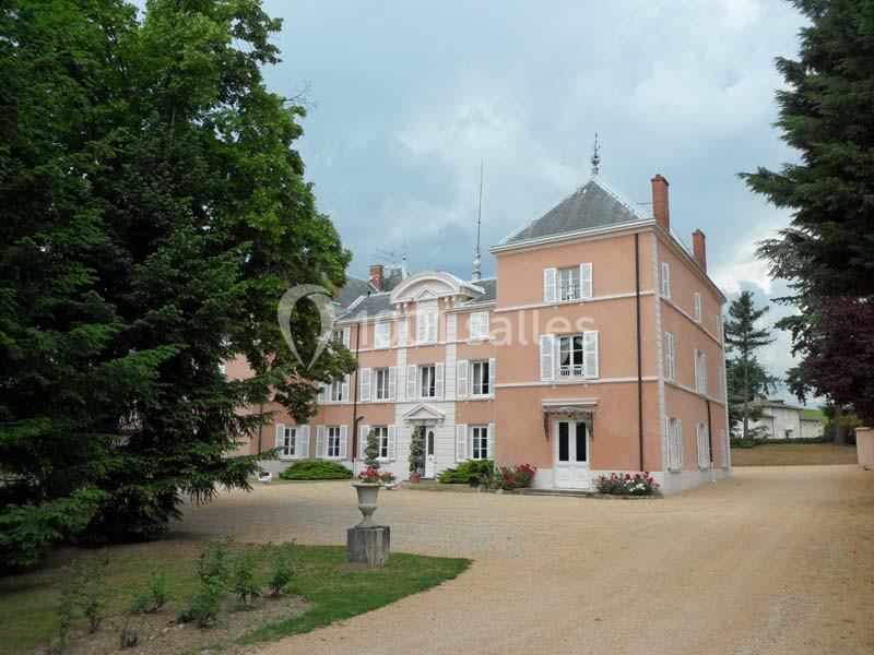 Manoir rose à trois étages entouré d'arbres, avec une allée en gravier et un ciel nuageux en arrière-plan.