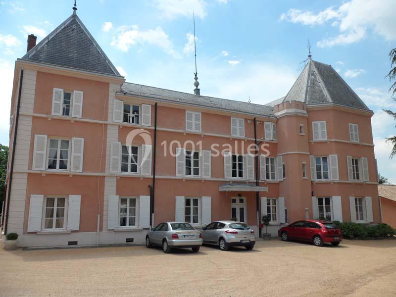 Façade d'un bâtiment rose à trois étages avec volets blancs, entouré de voitures stationnées sur une cour en gravier.