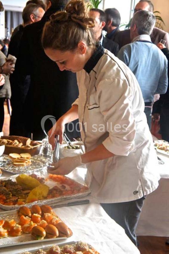 Une cheffe en tenue blanche prépare des plats sur un buffet lors d'un événement avec des convives en arrière-plan.