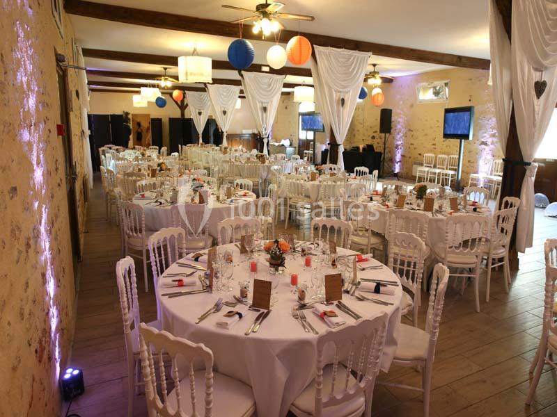 Salle de réception décorée avec des tables rondes dressées, chaises blanches et lanternes suspendues.