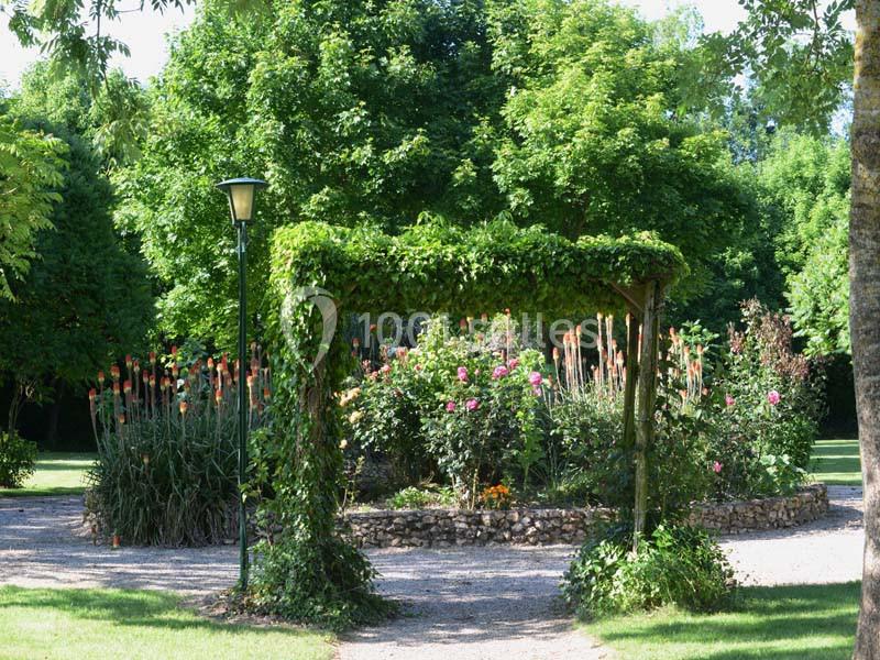 Allée de jardin avec une arche végétalisée, entourée de massifs fleuris et d'arbres verdoyants.