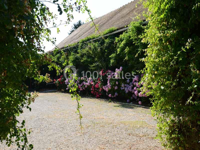 Cour bordée de végétation luxuriante et de fleurs colorées devant un bâtiment au toit de chaume.