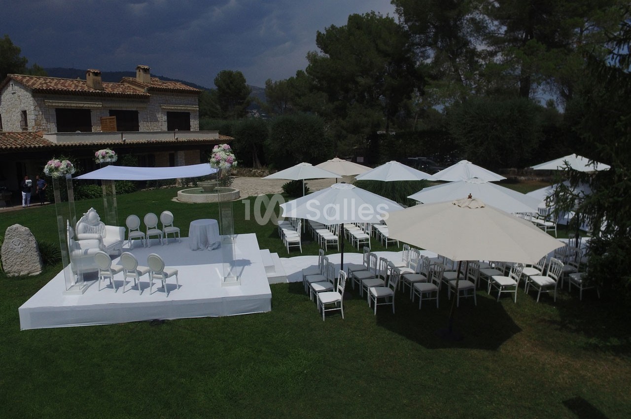 Cérémonie en plein air avec chaises blanches, parasols et décoration florale, devant une maison en pierre.