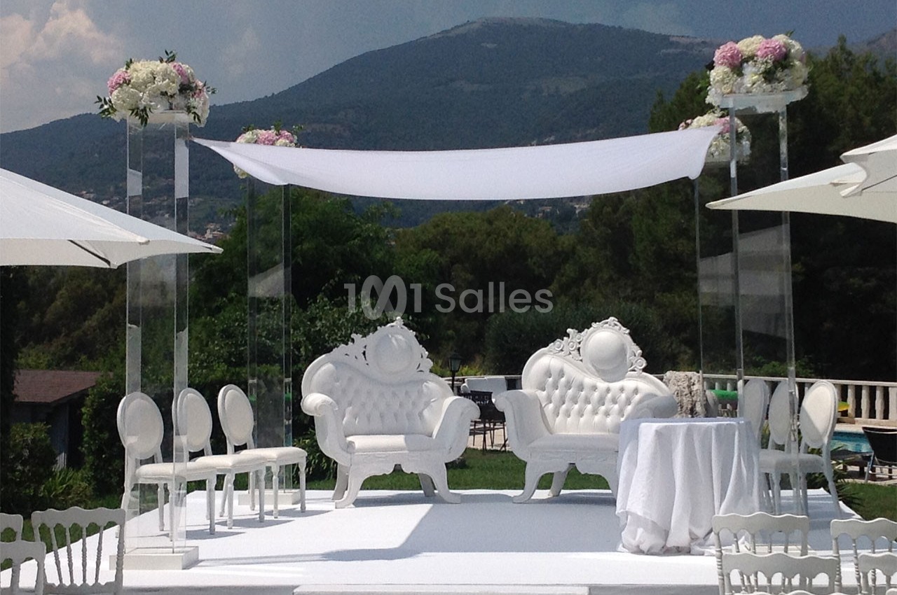 Décor de cérémonie en extérieur avec fauteuils blancs ornés, arche florale et vue sur des collines verdoyantes.