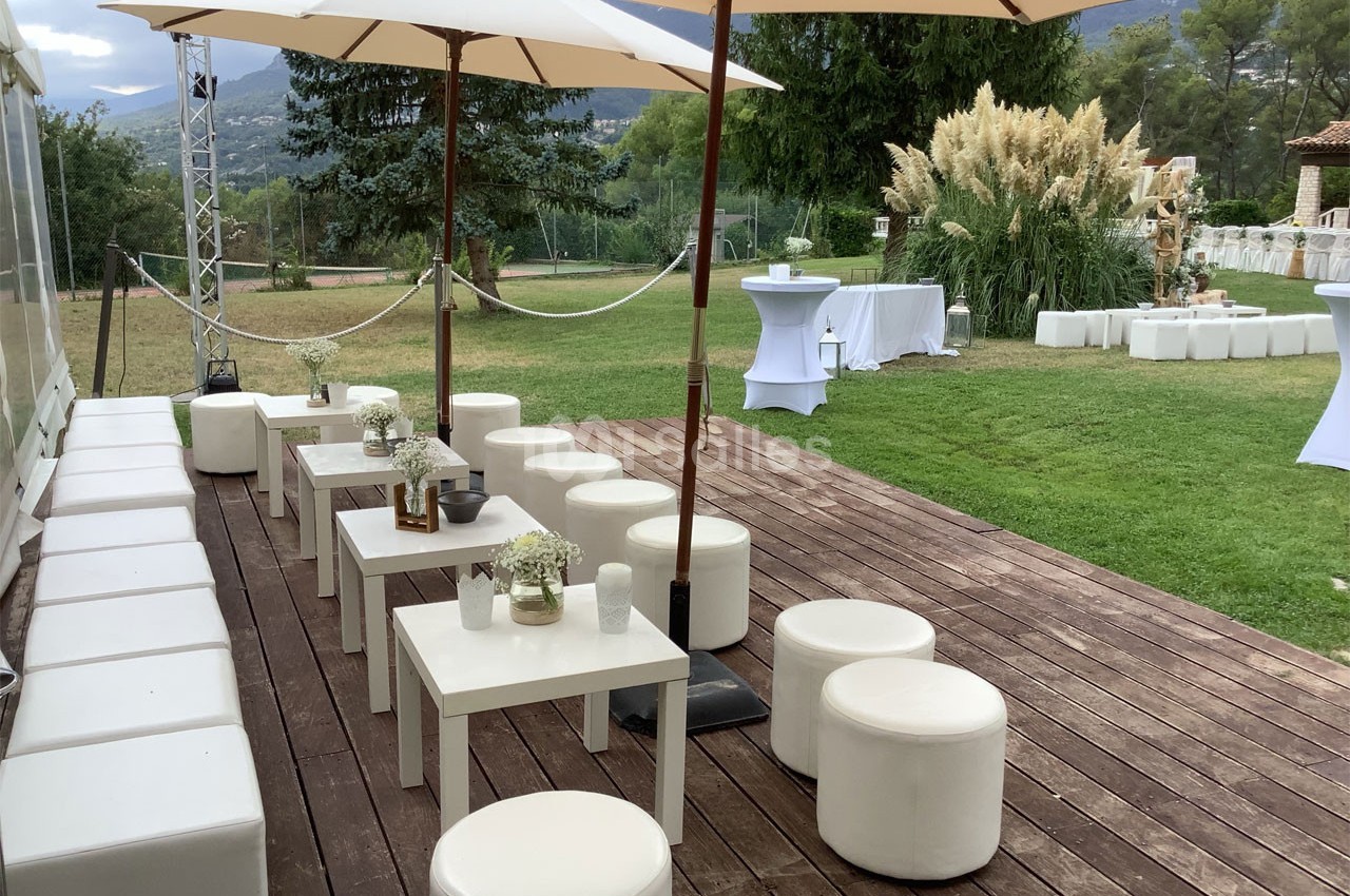 Terrasse en bois aménagée avec tables et sièges blancs, parasols et vue sur un jardin verdoyant.