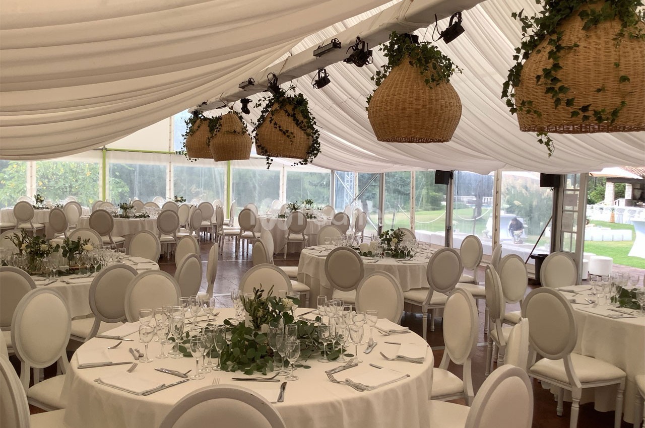 Salle de réception sous chapiteau avec tables rondes dressées, chaises blanches et suspensions en osier décorées de verdure.