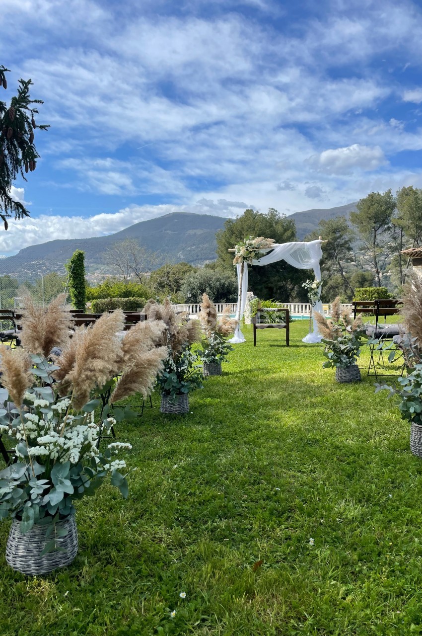 Allée décorée de fleurs et de pampas menant à une arche de mariage en extérieur, avec vue sur des collines verdoyantes.