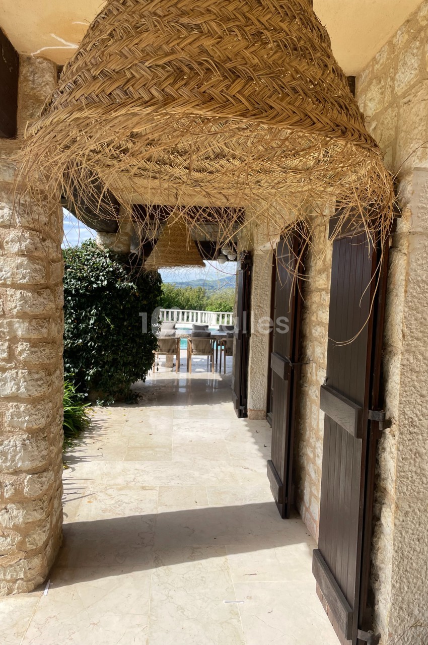 Passage extérieur en pierre avec portes en bois, grande suspension en osier et vue sur une terrasse ensoleillée.