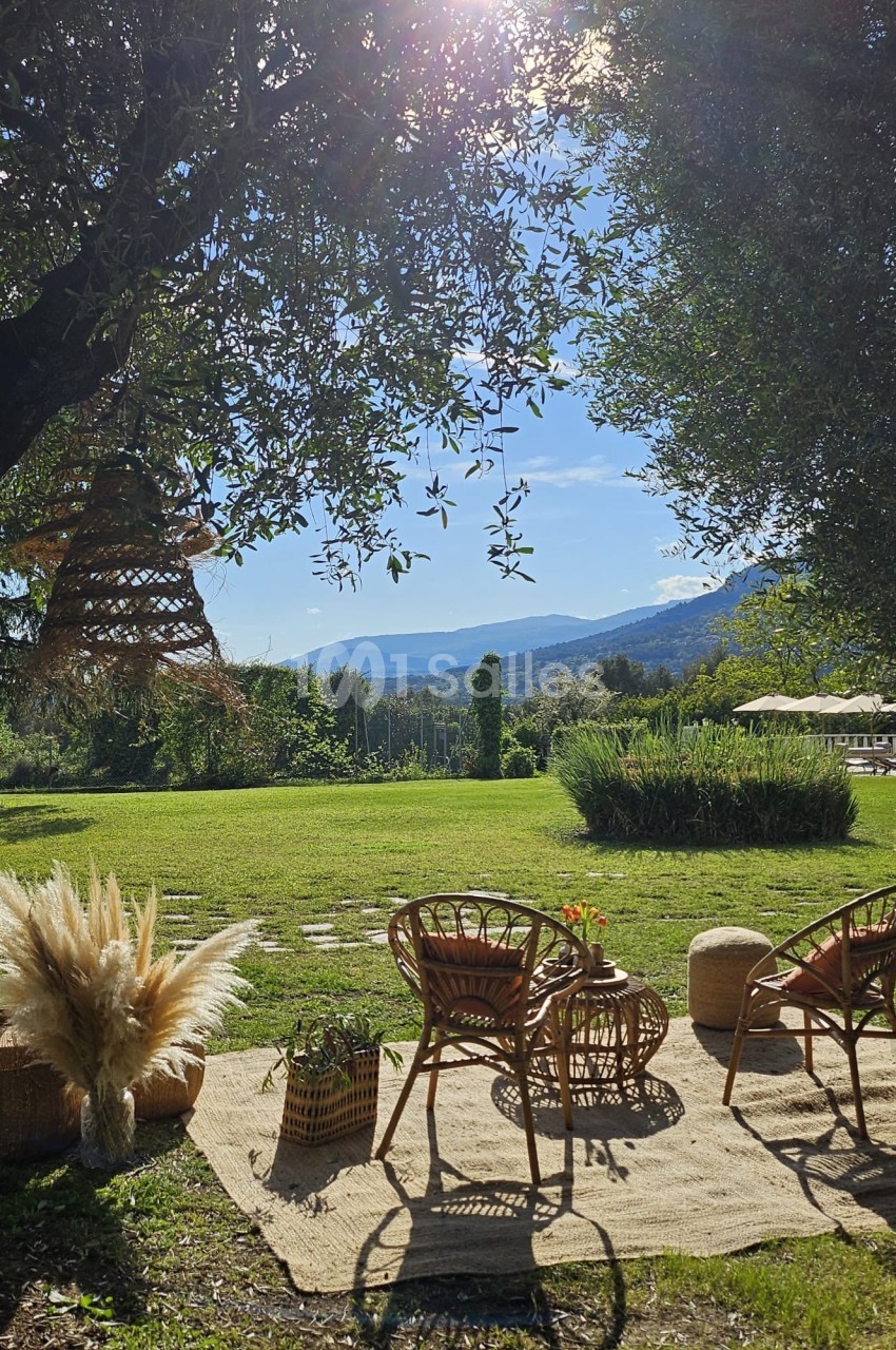 Espace extérieur avec fauteuils en rotin, tapis, plantes et vue sur un jardin verdoyant sous un ciel ensoleillé.