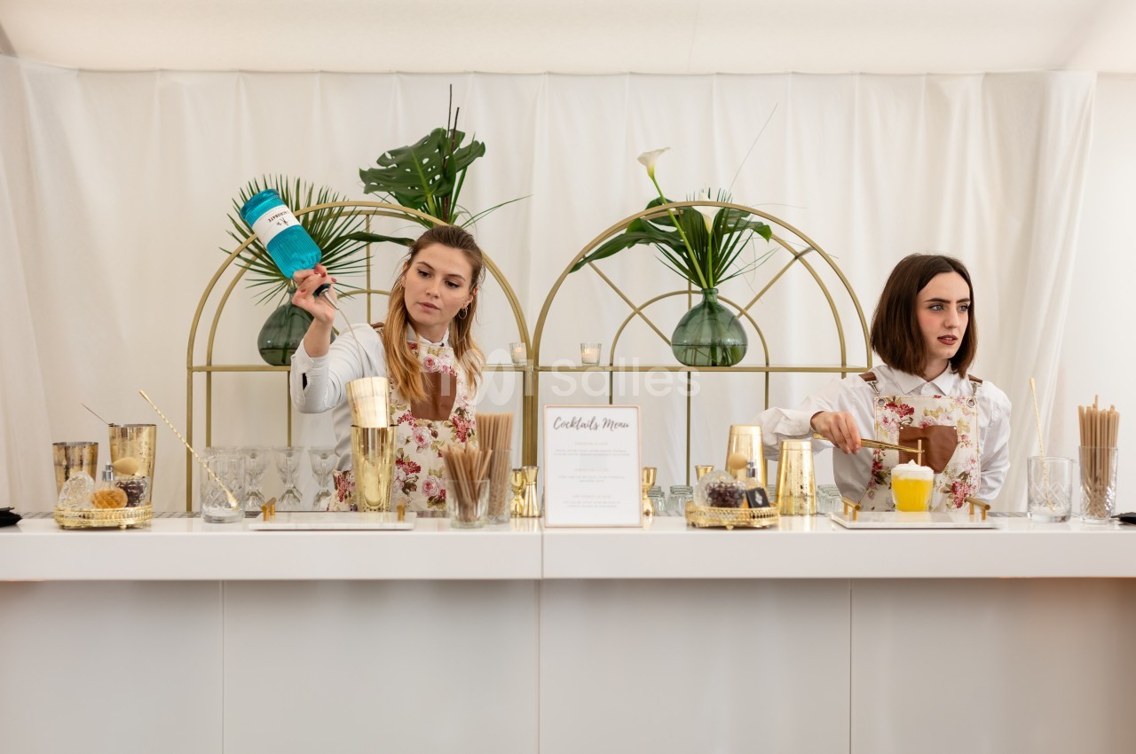 Deux femmes préparent des cocktails derrière un comptoir blanc décoré de plantes et d'accessoires dorés.