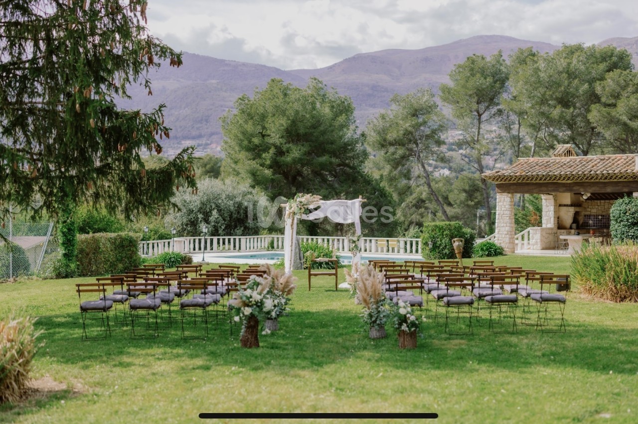 Chaises alignées devant une arche décorée de fleurs dans un jardin verdoyant avec des montagnes en arrière-plan.