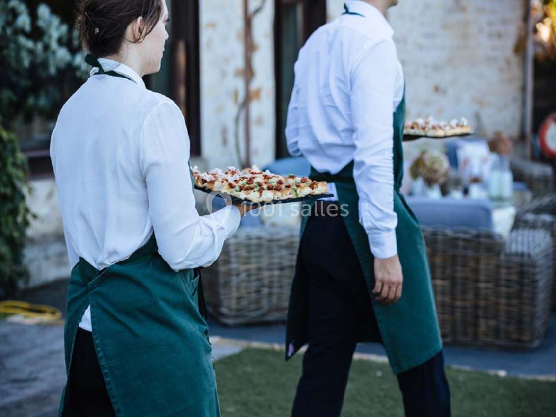 Deux serveurs en tablier vert portent des plateaux de nourriture dans un espace extérieur près de tables dressées.