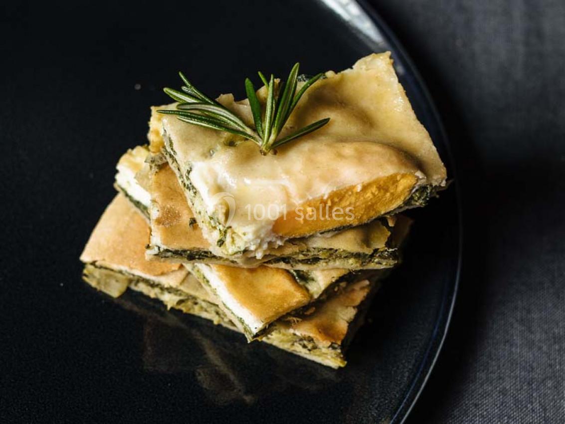 Tranches de quiche aux légumes et fromage empilées sur une assiette noire, garnies d'une branche de romarin.