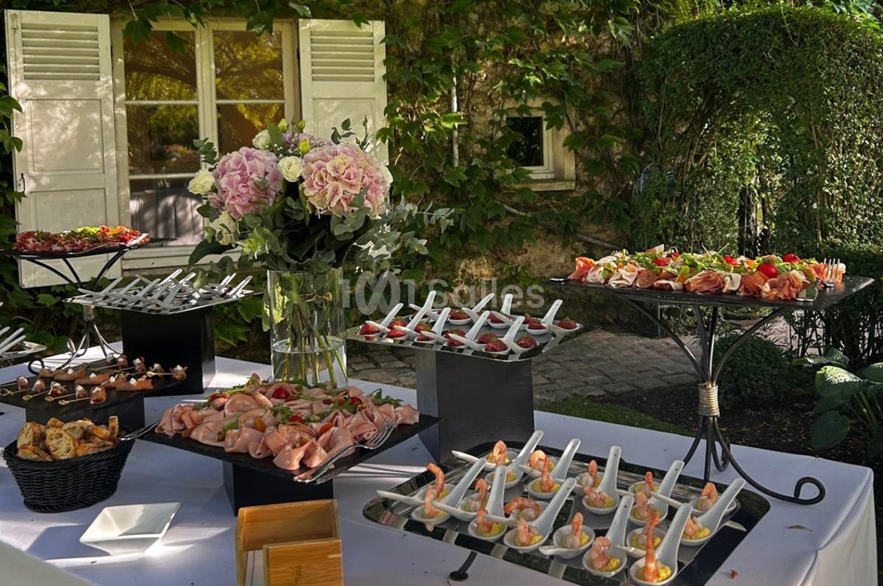 Buffet en extérieur avec divers plats et amuse-bouches disposés sur des présentoirs, entouré de verdure et de fleurs.